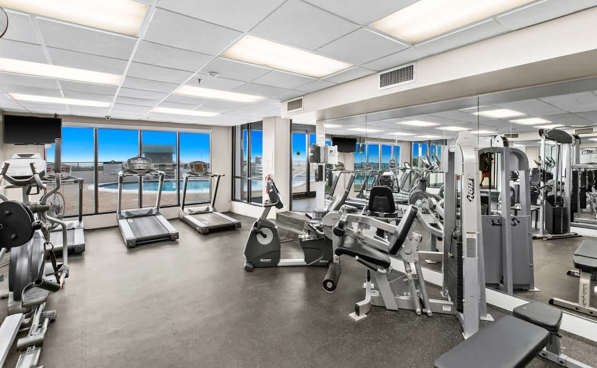 A bright fitness room with a range of modern exercise equipment, including weight machines and cardio equipment, set against a mirrored wall and large windows overlooking the exterior. The floor is dark, durable material.
