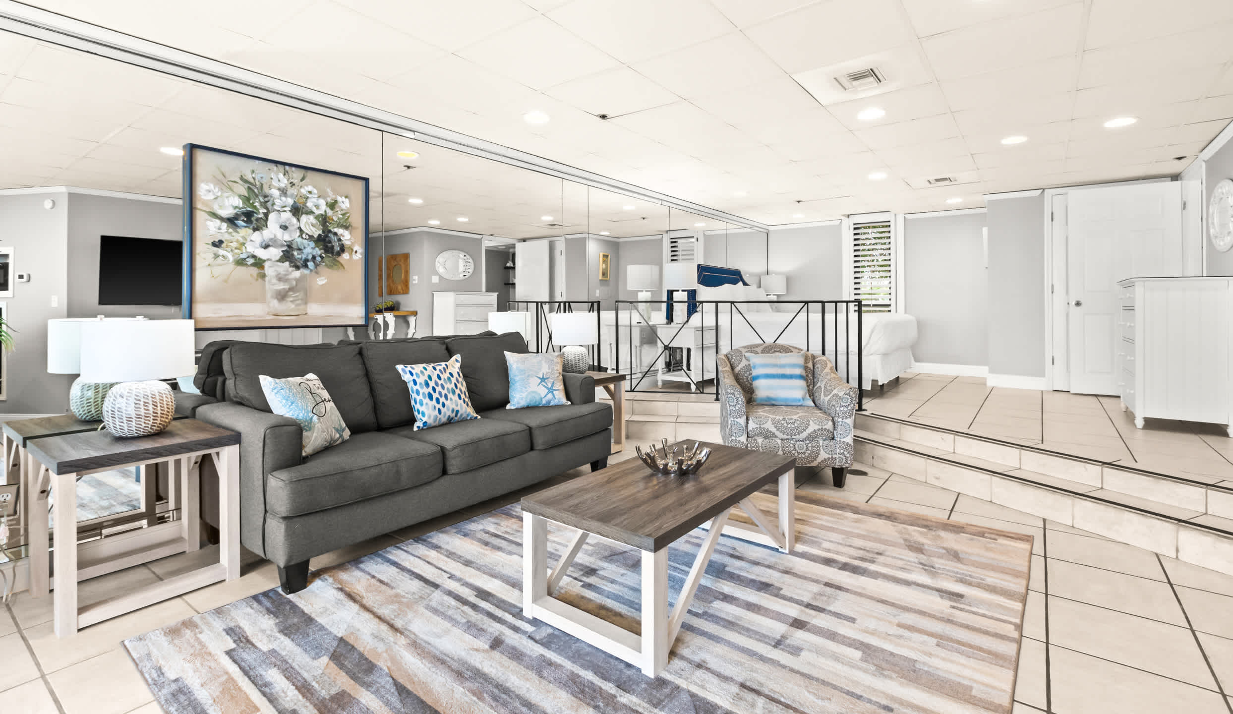 A spacious living room with a dark gray sofa, a patterned area rug, and a light wood coffee table, featuring a large mirror wall reflecting a painting and a partial view of a bedroom area beyond.