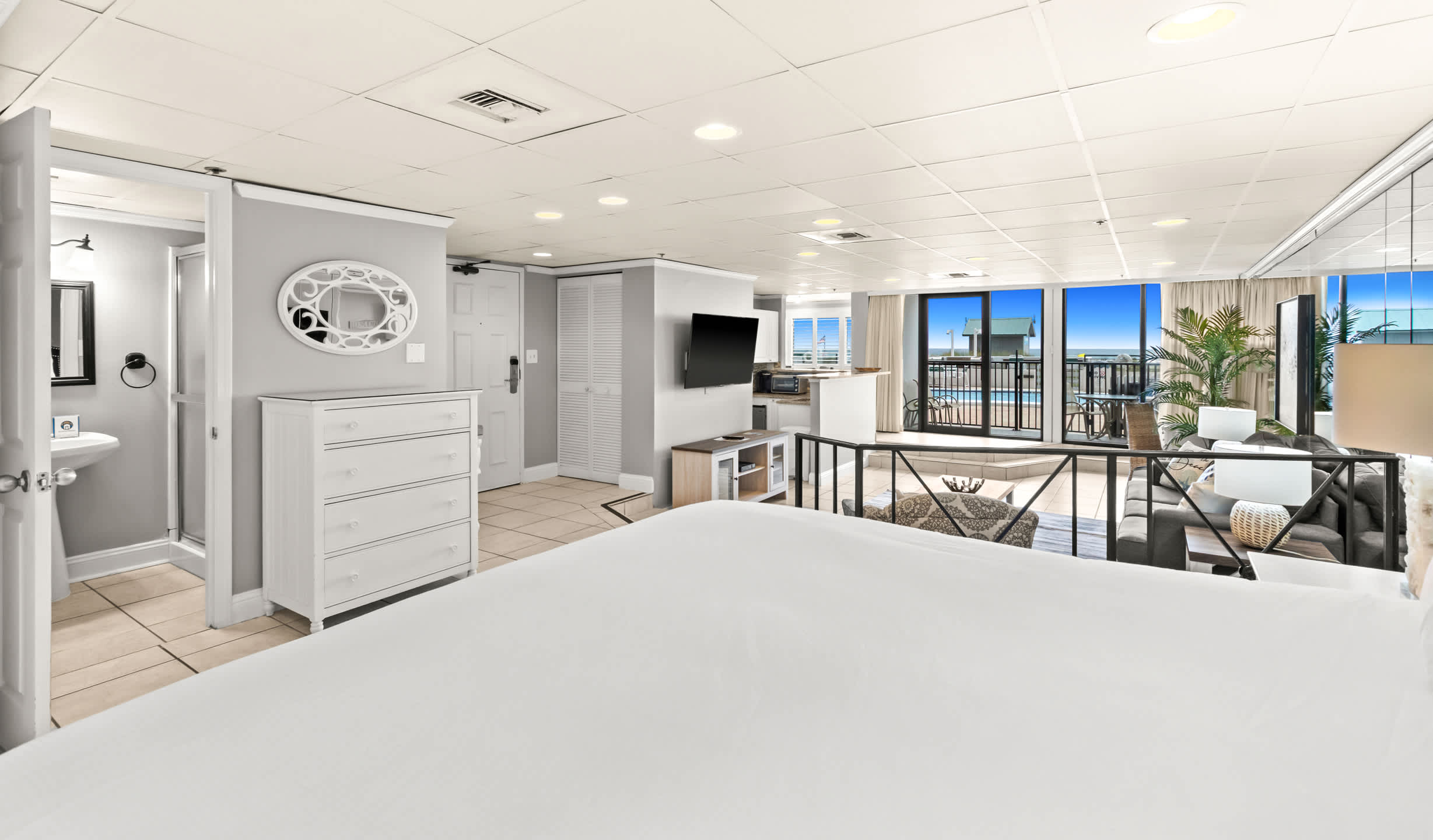 View from the king bed with a blue headboard, looking towards the living area with a dresser and a wall-mounted TV, and a balcony with ocean views in the distance. A bathroom door is visible on the left.