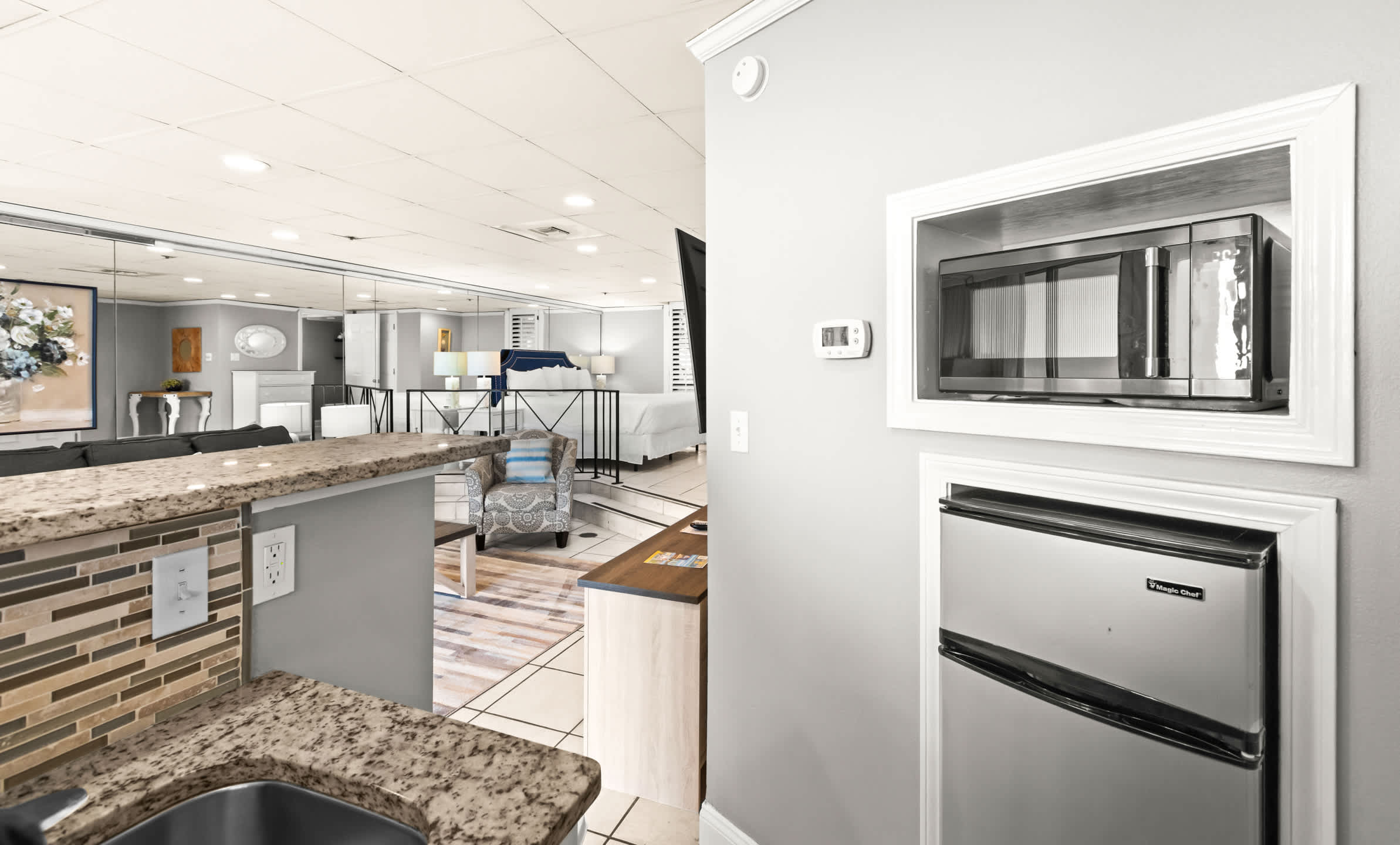 A view of the kitchenette area from the side, showing a built-in microwave and a mini-fridge below, with a partial view of the living room and mirrored wall in the background.