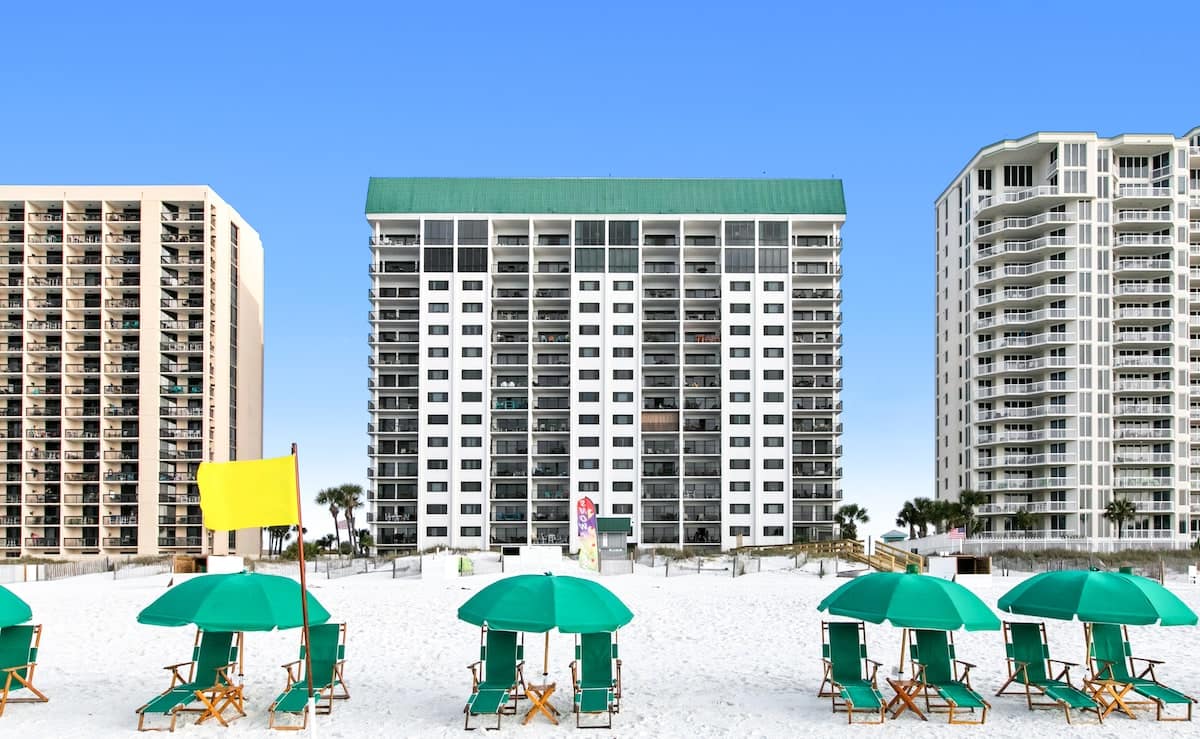 Full exterior view of a tall, white condominium building with a green roof, situated directly on a wide white sand beach with rows of green beach umbrellas and chairs in the foreground.