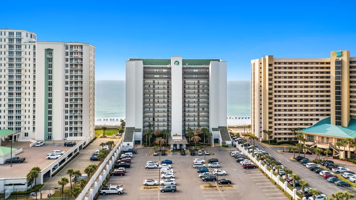 An aerial view showcasing the front facade of a tall condominium building with a green roof, surrounded by a large parking lot and other high-rise structures. The ocean is visible in the background.