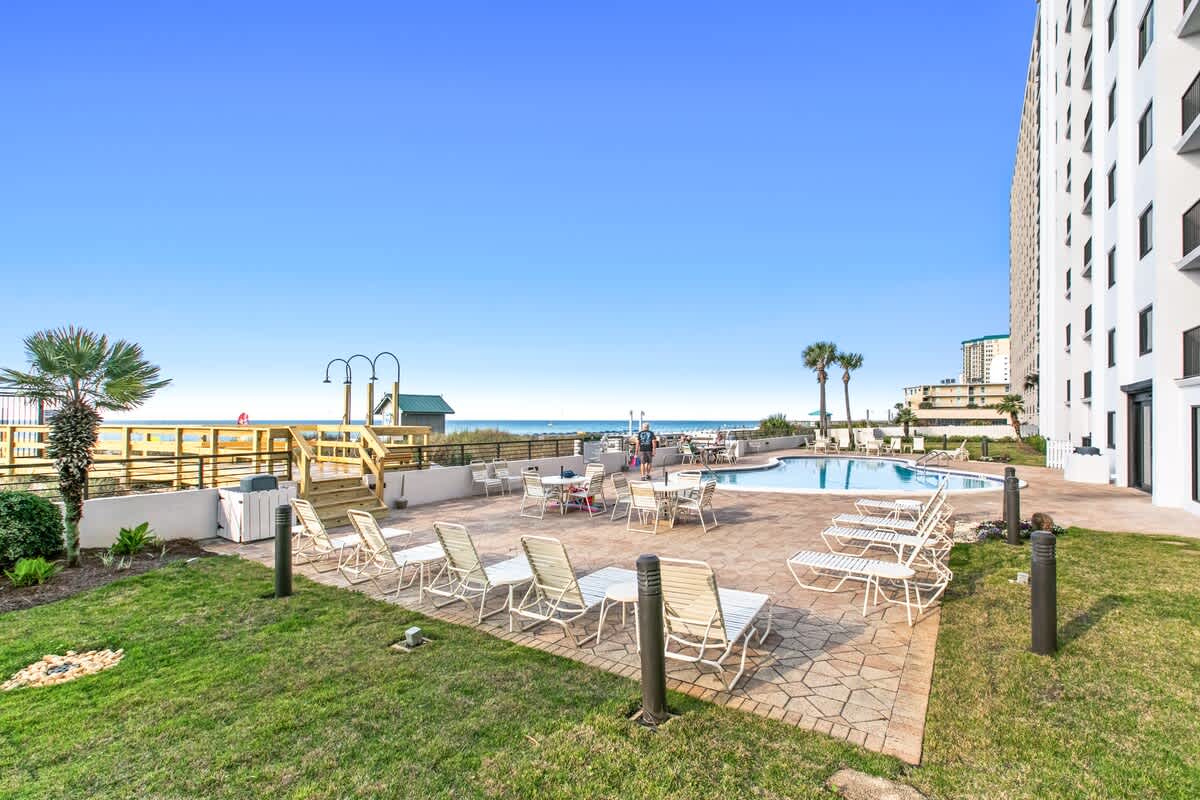 Outdoor communal swimming pool area with lounge chairs and tables, surrounded by green grass and paving, offering a distant view of the ocean and beach access, with a tall building in the background.