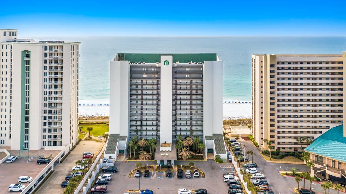 A direct aerial view of the central high-rise condominium building, featuring a distinctive green roof and multiple balconies, flanked by a spacious parking area and other tall structures. The ocean is clearly visible in the distance.