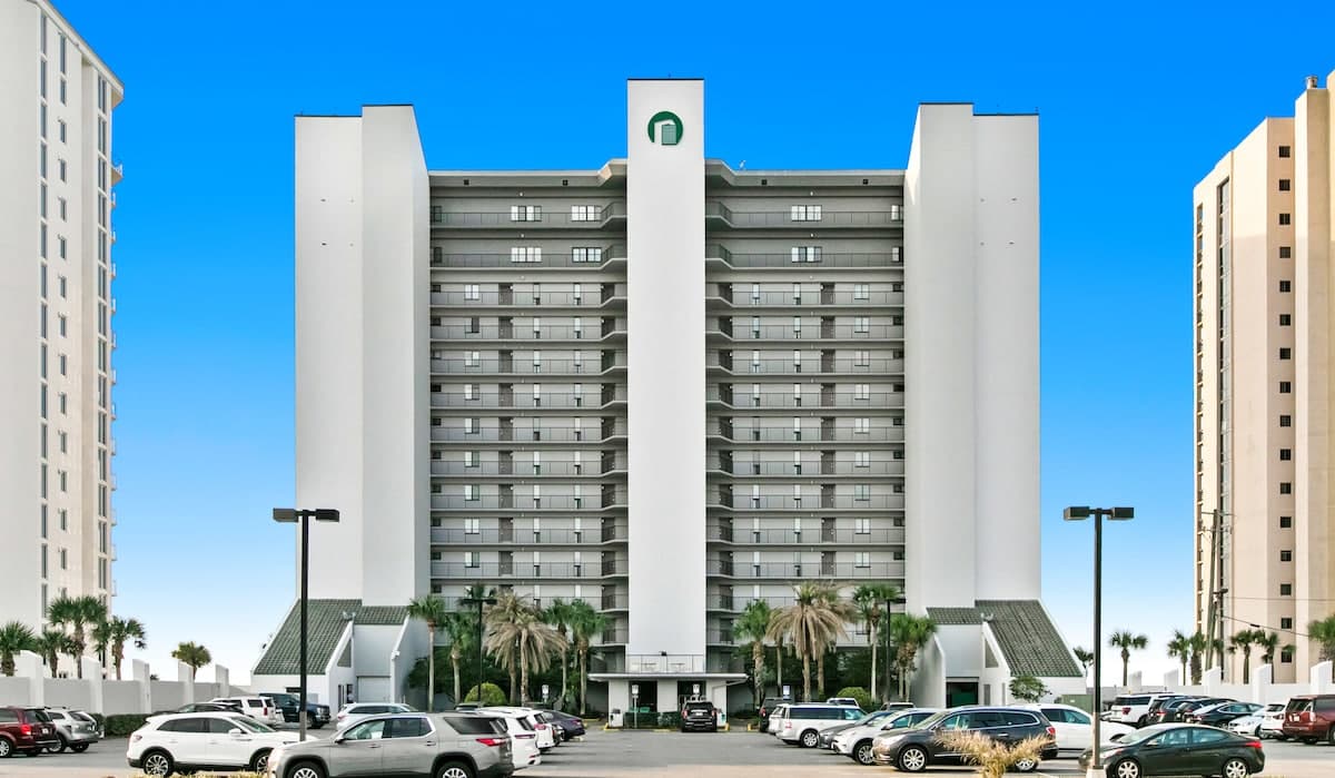 Symmetrical front view of a multi-story white condominium building with a prominent green logo, featuring numerous balconies and a large parking area with vehicles, all set against a clear blue sky.