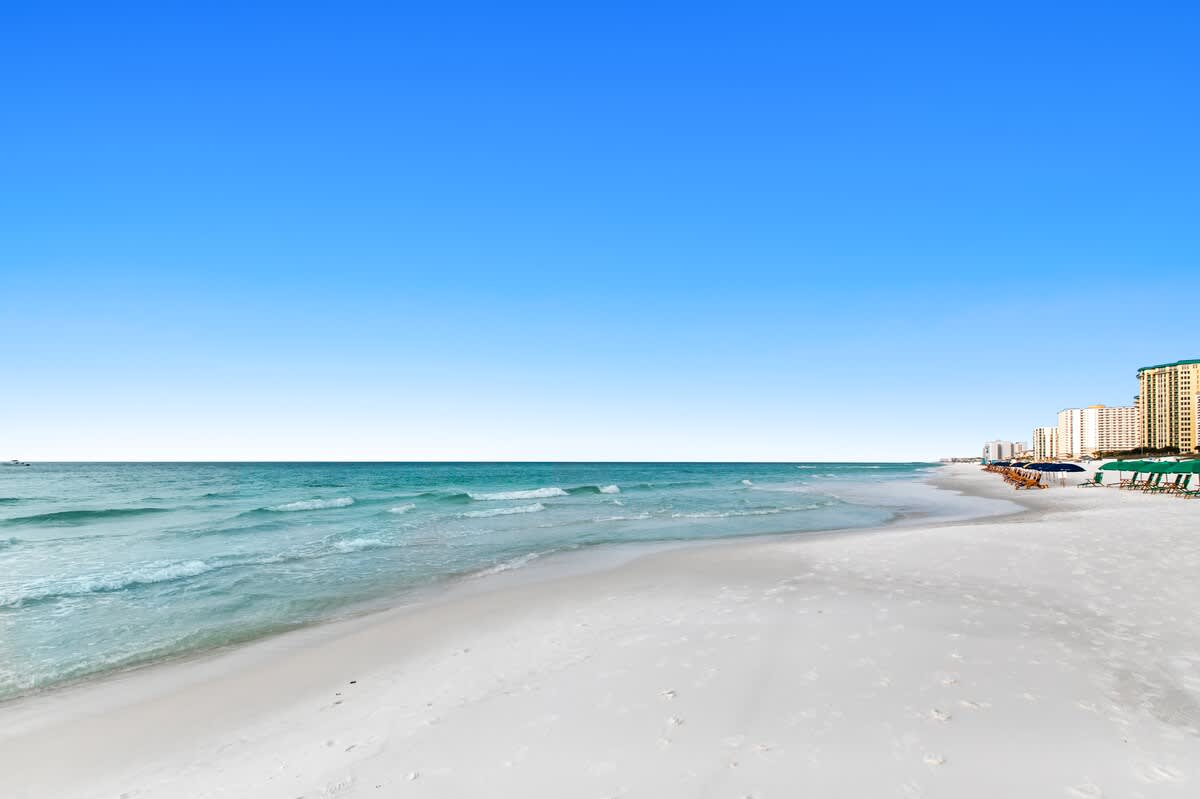Panoramic view of a long, wide white sand beach with gentle waves, extending along a coastline dotted with high-rise condominium buildings under a clear blue sky.