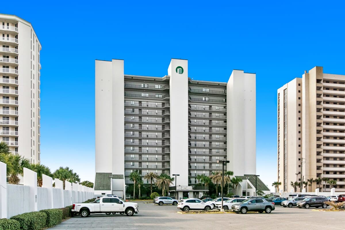 A tall, modern white condominium building with a distinctive green logo at the top, surrounded by a large parking lot filled with cars. Palm trees and other buildings are visible under a bright blue sky.