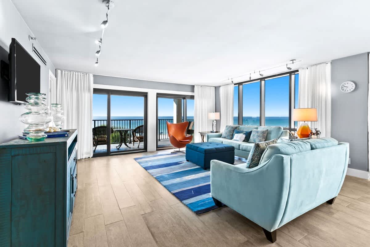 Bright living room with light blue sofas, an orange accent chair, and a striped blue rug, all arranged to maximize the stunning panoramic ocean views through floor-to-ceiling windows and balcony access. A teal console table holds a TV.
