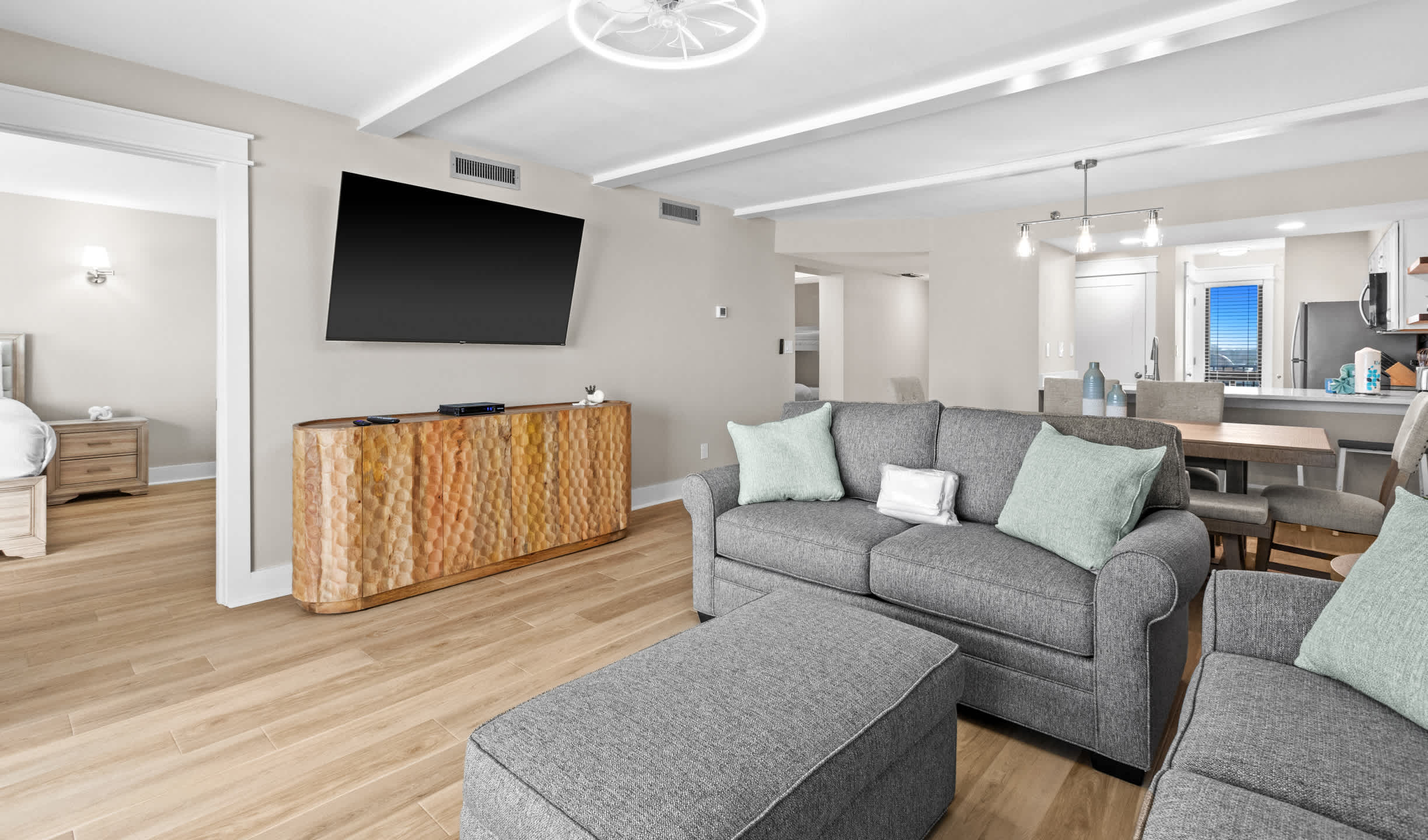 Modern living room with light wood-look flooring, a gray sofa, and a large wall-mounted TV above a textured wooden console. A doorway leads to a bedroom on the left.