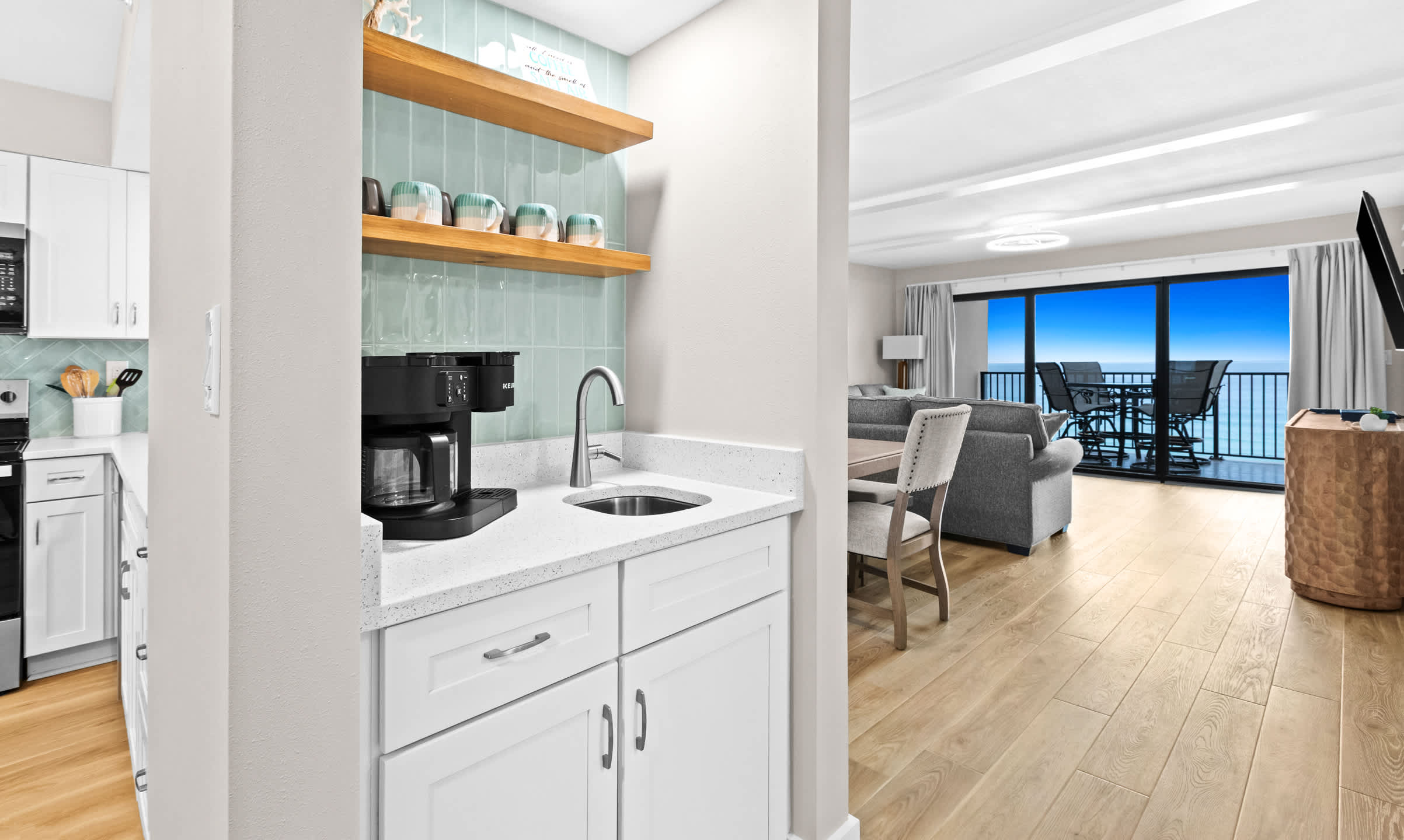 Dedicated wet bar or coffee station area with white shaker cabinets, a small sink, and open wooden shelves displaying mugs, set against a light blue herringbone tile backsplash.