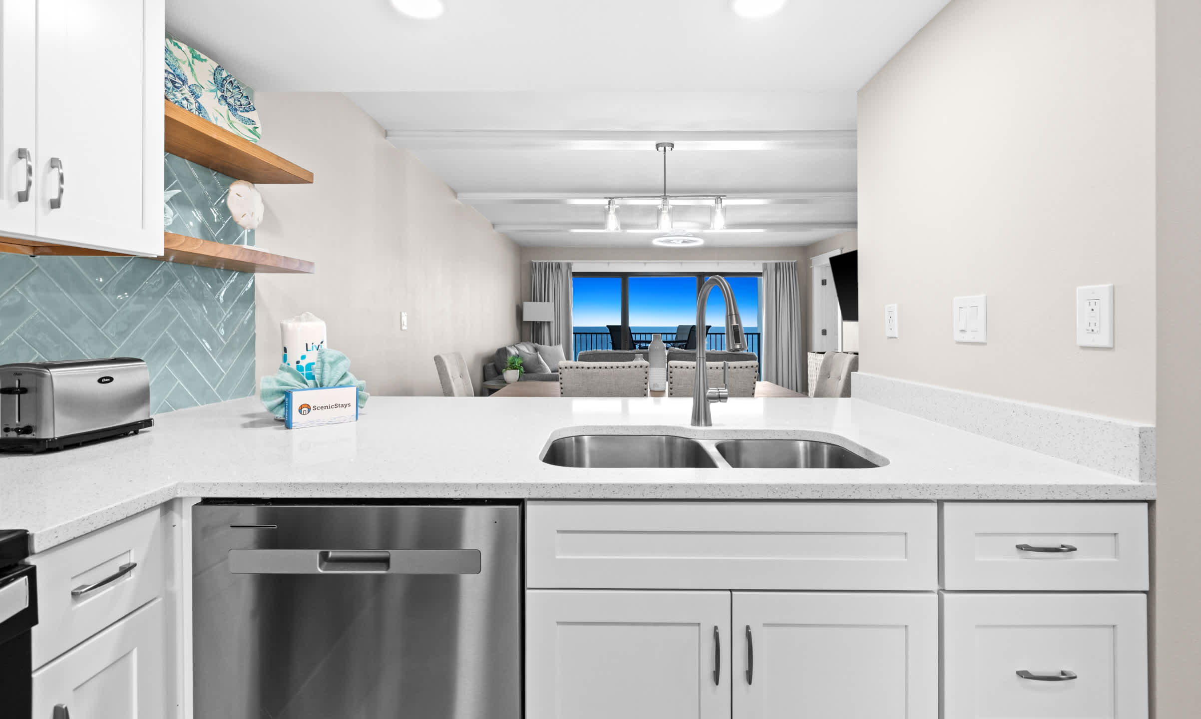 View from the kitchen sink area, showcasing a double stainless steel sink, white quartz countertops, and a stainless steel dishwasher, with an open view towards the living room and ocean beyond.