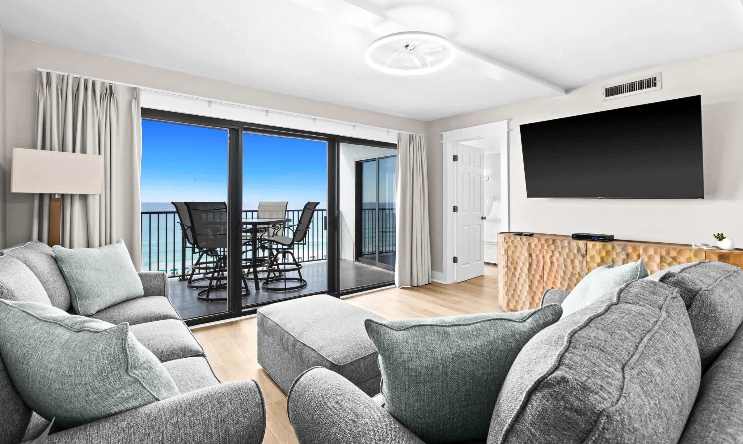 Bright living room with gray sofas and an ottoman, featuring a large sliding glass door leading to a balcony with outdoor seating and panoramic ocean views under a clear blue sky.