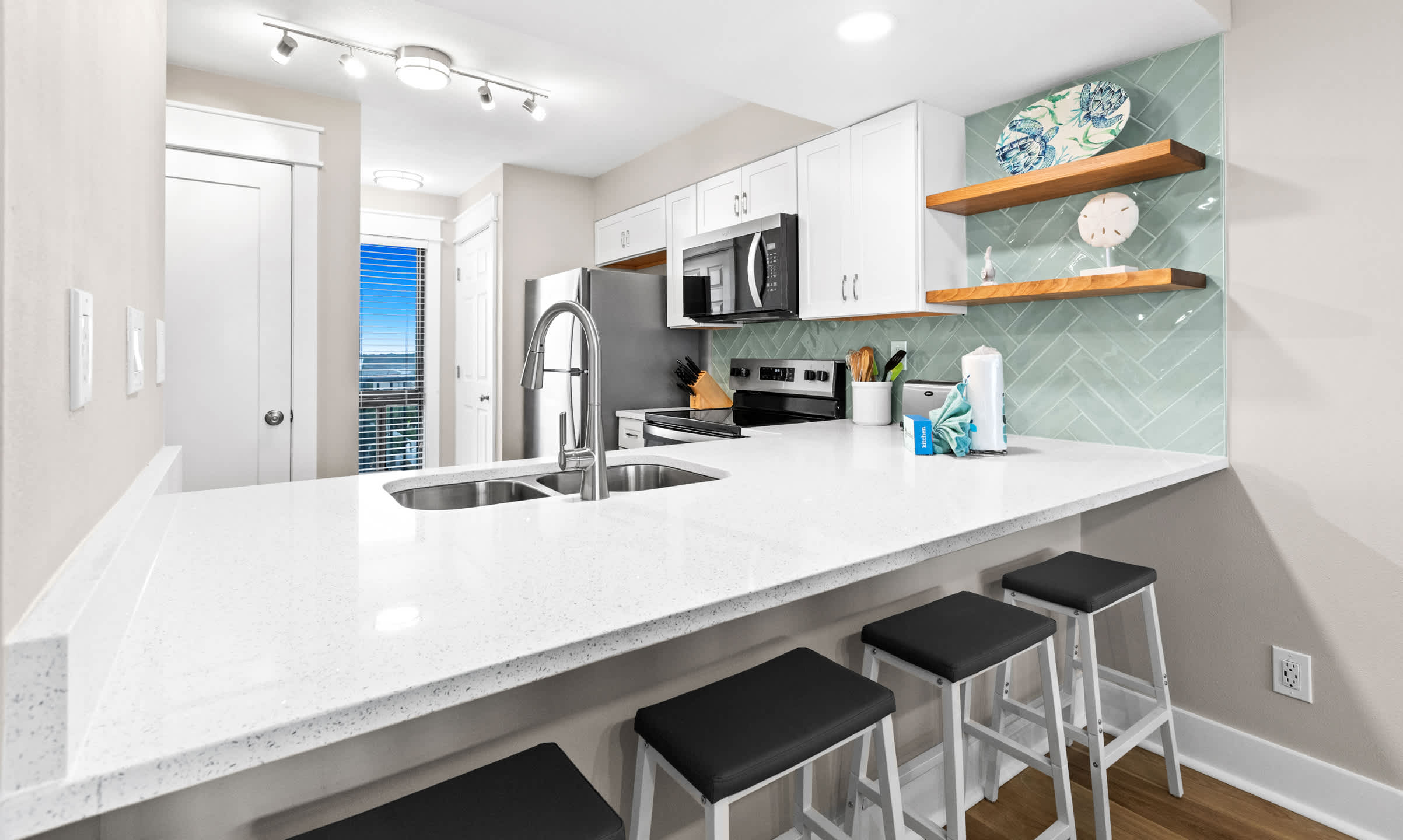 Spacious kitchen island with white quartz countertops and three black bar stools, offering a breakfast bar, and an open view of the modern kitchen with stainless steel appliances and light wood-look flooring.
