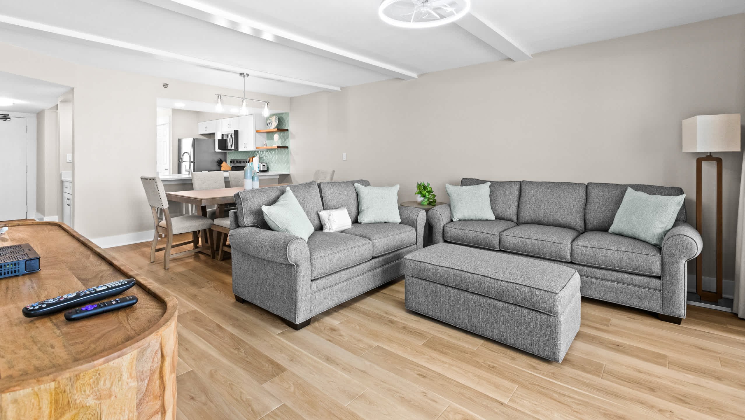 Bright living room featuring light wood-look flooring, two gray sofas with light blue throw pillows, and a matching ottoman. A dining area and kitchen are visible in the background.