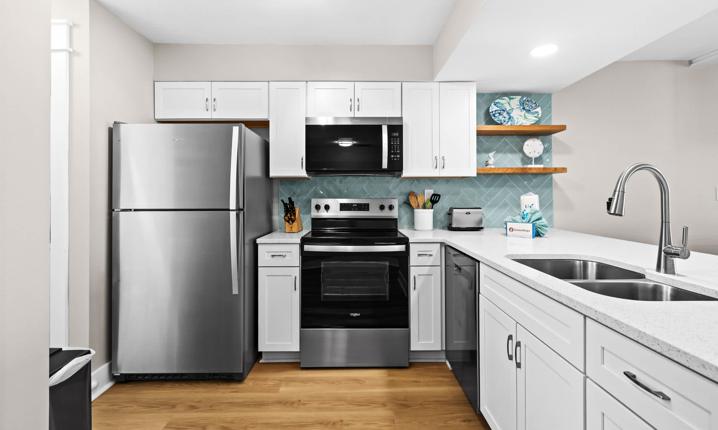Close-up of the modern kitchen featuring white shaker cabinets, stainless steel refrigerator, oven, and microwave, with a light blue herringbone tile backsplash and open wooden shelving.