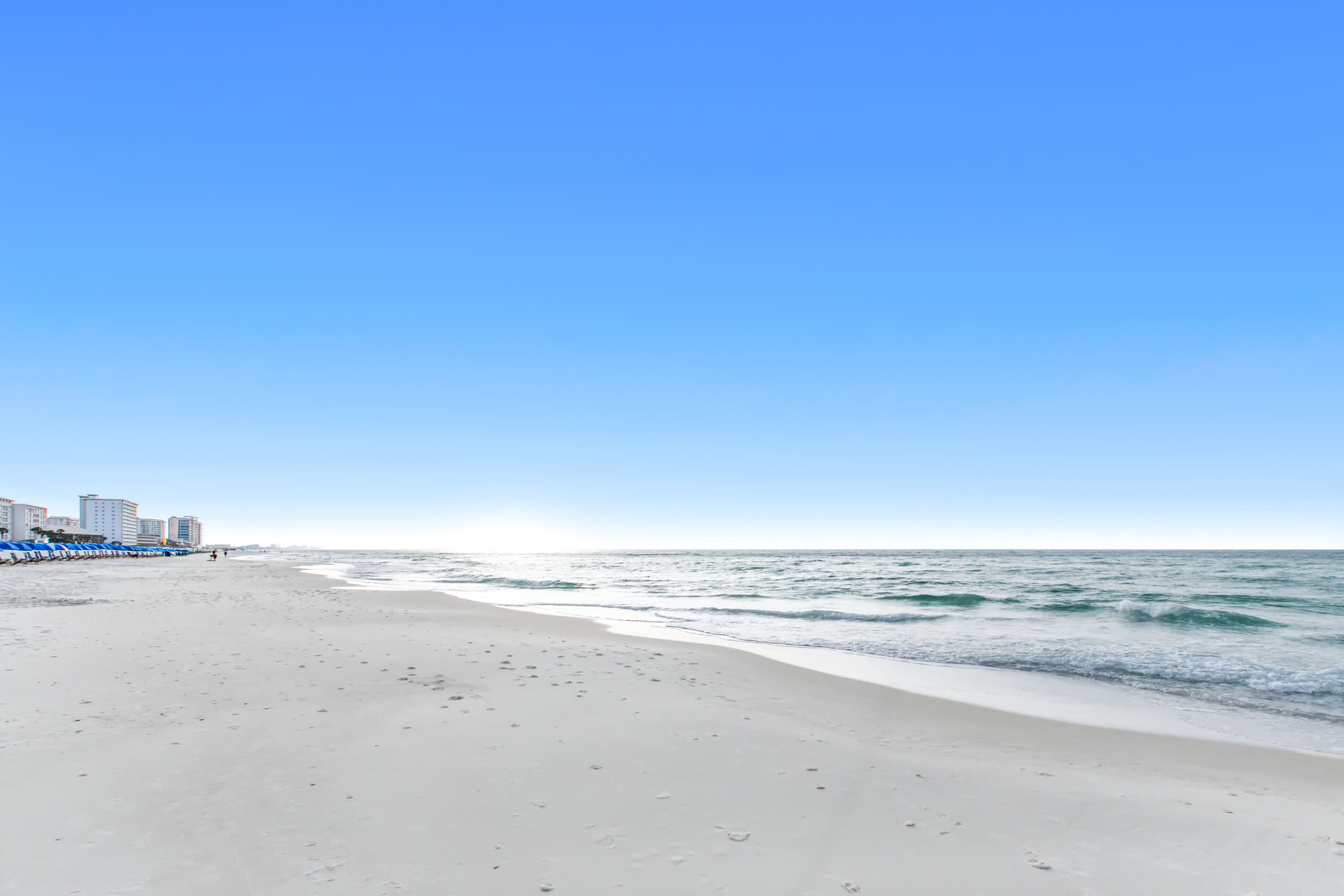 Serene ground-level view of a pristine white sand beach meeting the gentle turquoise waves of the ocean under a clear blue sky. Distant buildings line the horizon.