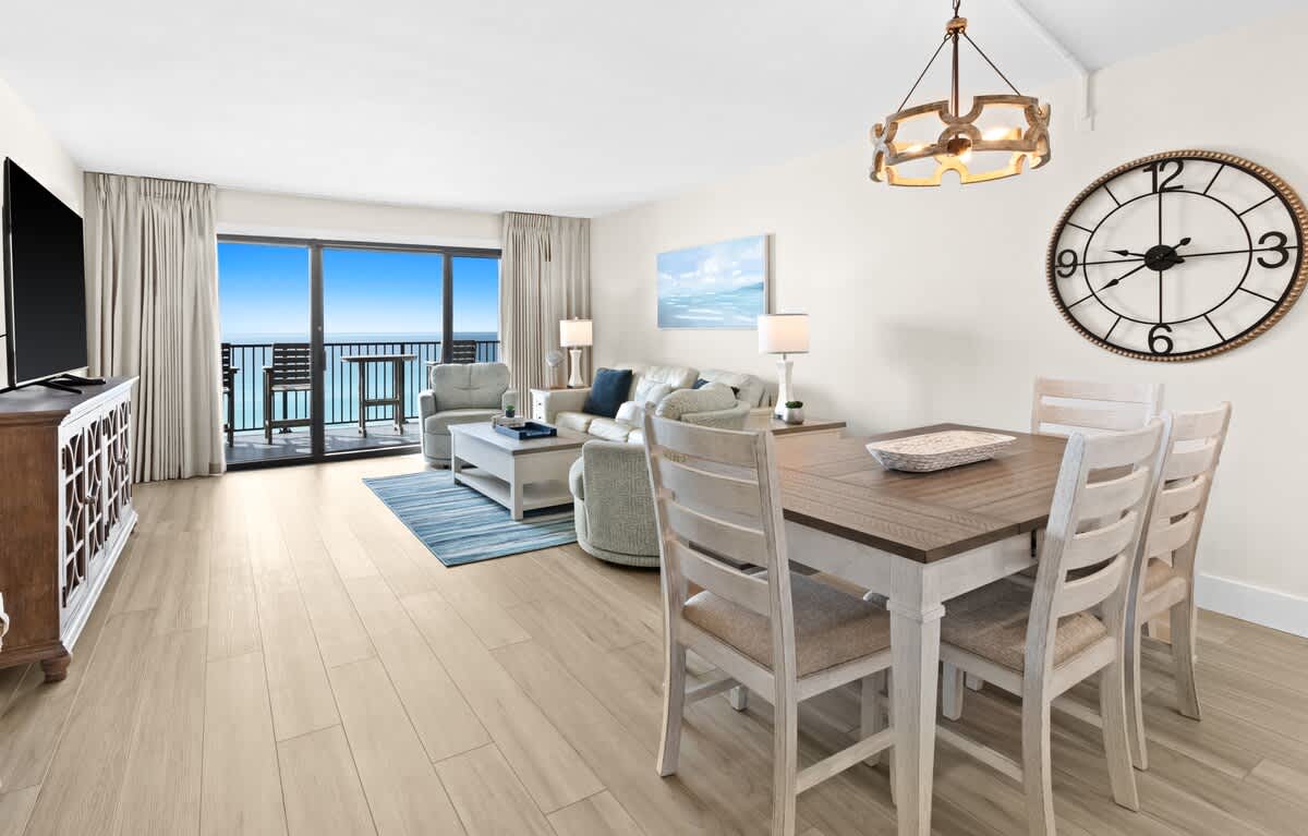 Bright and airy open-concept living and dining area with light wood-look flooring and white walls. The living space features a light-colored sofa and armchairs, while the dining area has a wooden table with seating for four, all overlooking a balcony with ocean views.