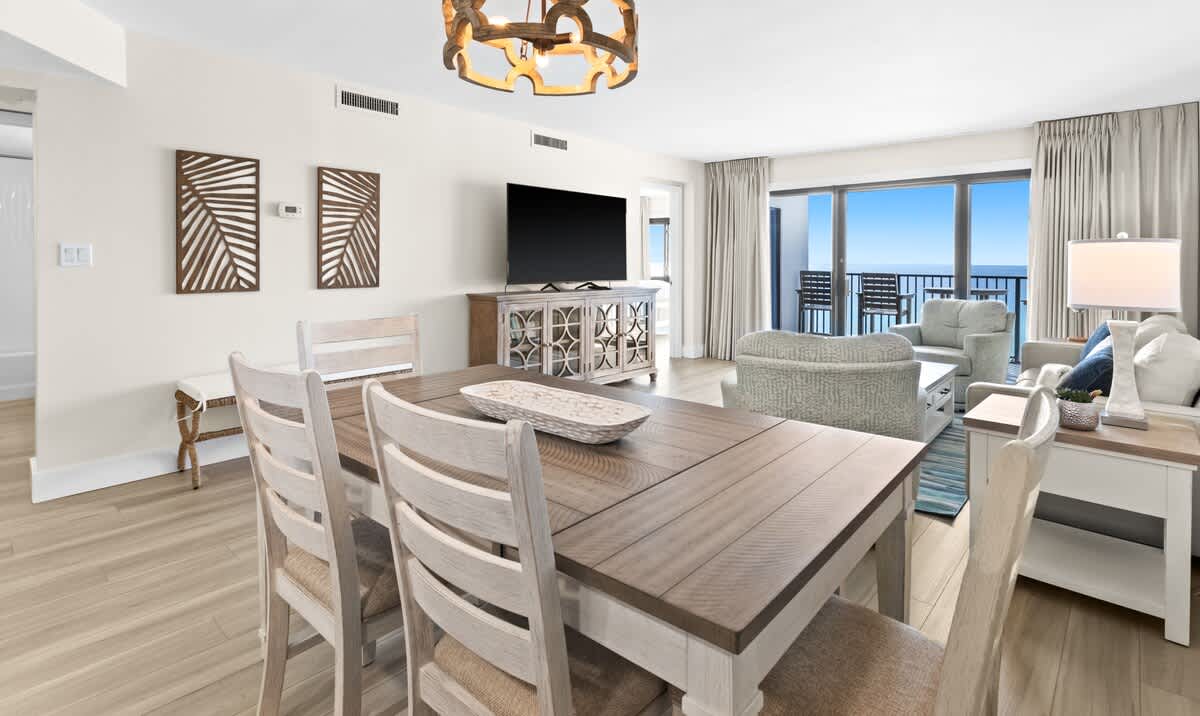 Spacious living and dining area with light wood-look flooring and a large flat-screen TV on a rustic wooden console. The dining table seats four, and the living area features comfortable seating, all bathed in natural light from the balcony overlooking the ocean.