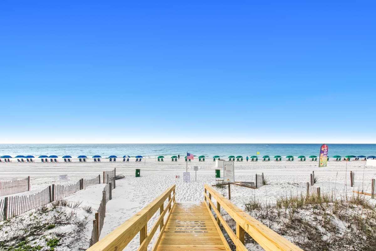 Wooden boardwalk providing direct access to a wide, white sandy beach lined with rows of green beach umbrellas and chairs. The clear blue ocean stretches into the horizon under a bright sky.