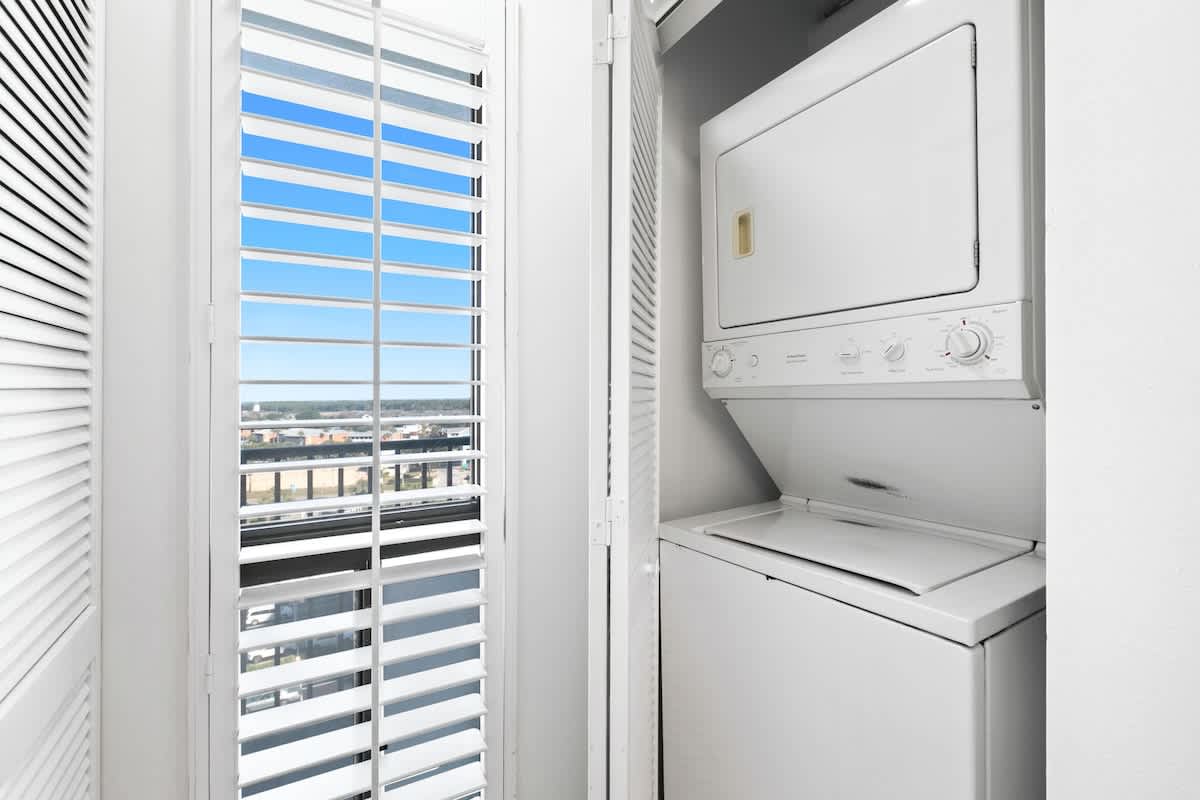Compact laundry closet featuring a stacked white washer and dryer unit, located next to a window with white plantation shutters.