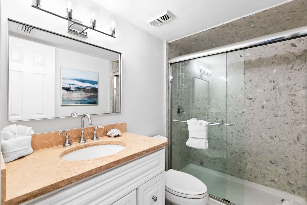 Modern bathroom featuring a light brown granite-like vanity with a white sink, chrome faucet, and a glass-enclosed shower with speckled gray tile walls. A large mirror and contemporary light fixture are above the vanity.