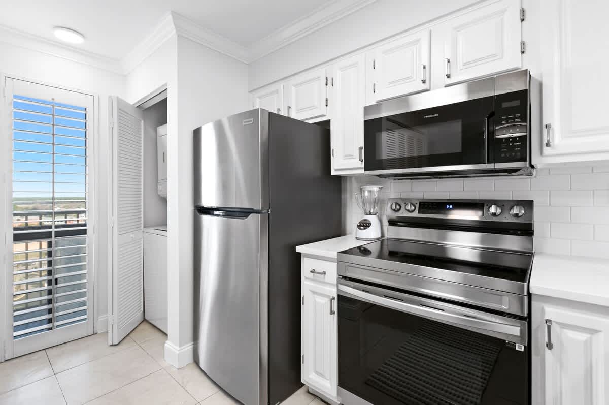 A well-equipped kitchen features white shaker cabinets, stainless steel refrigerator, oven, and microwave, with a white subway tile backsplash and a laundry closet visible in the background.