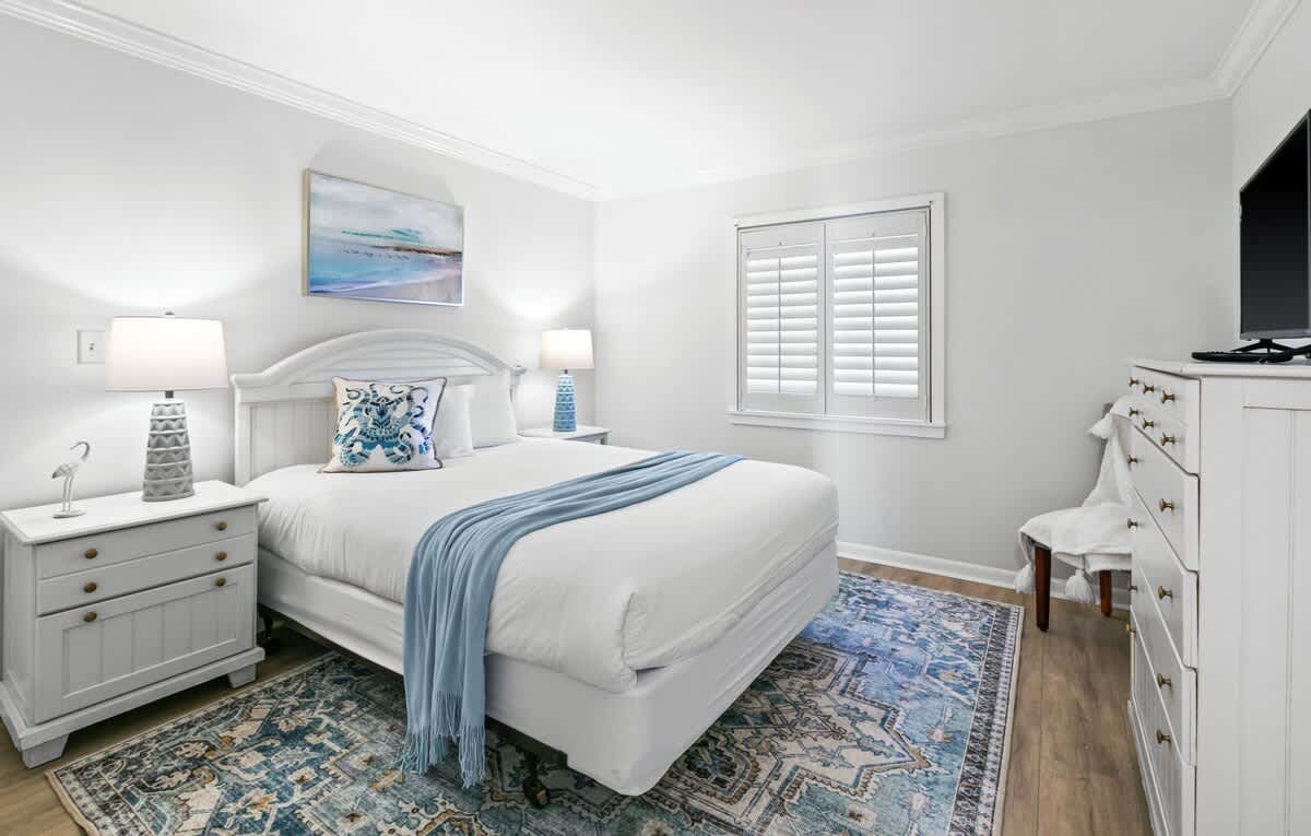 A cozy bedroom features a white queen-sized bed with a blue throw blanket and decorative pillows, flanked by two gray nightstands with lamps, and a white dresser with a TV, all on a patterned blue rug.
