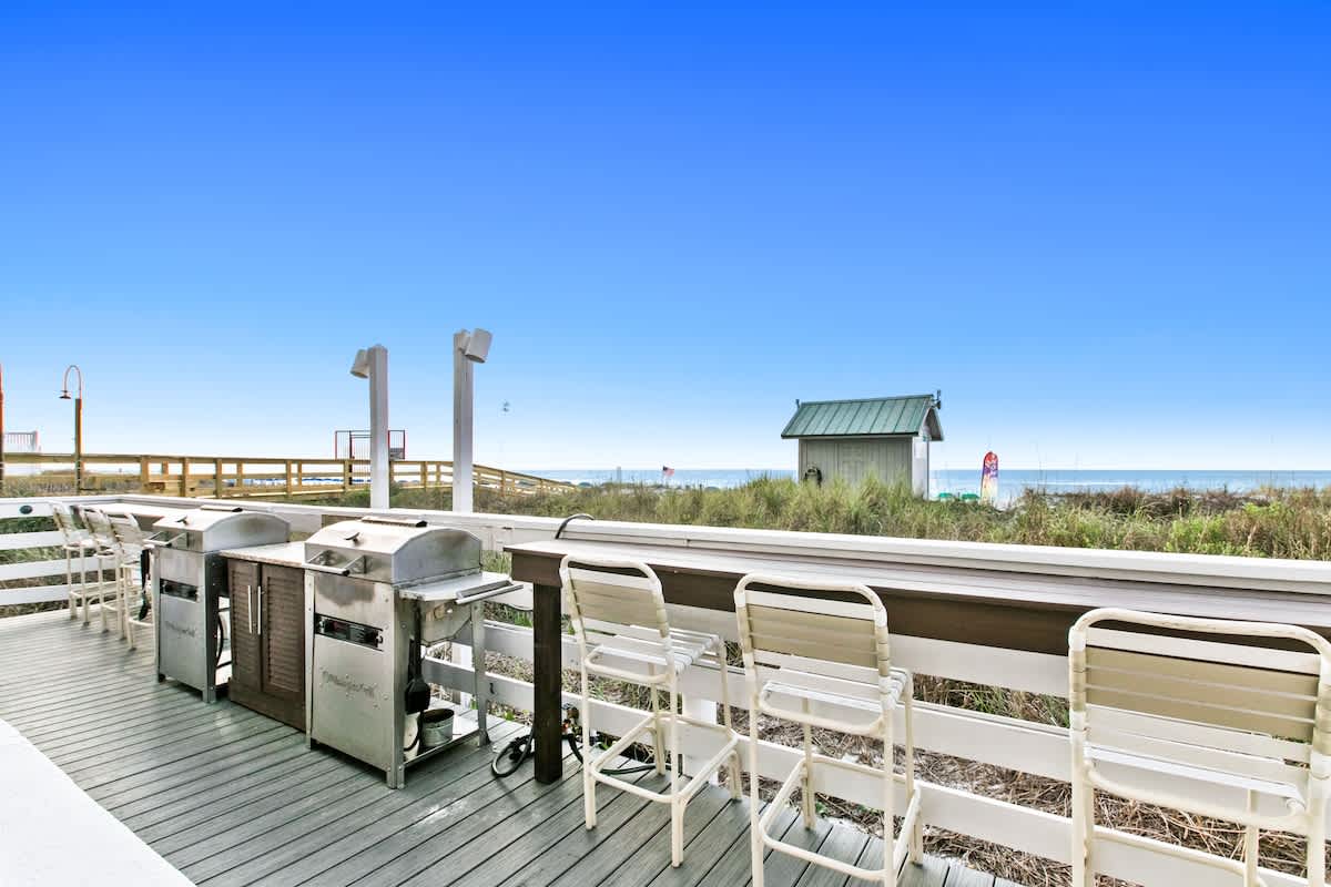 Outdoor grilling station with multiple stainless steel BBQ grills and a long bar counter with high chairs, situated on a wooden deck overlooking a grassy dune and the ocean.