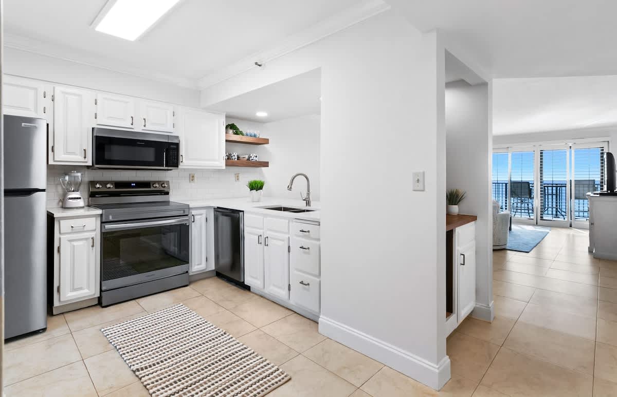 The bright kitchen features white shaker cabinets, stainless steel appliances including a refrigerator, oven, and microwave, white countertops, and a double sink, with a view towards the living area.