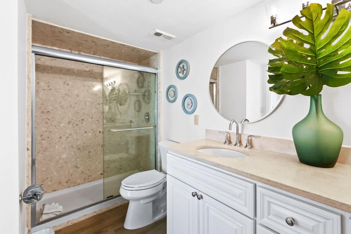 A bright bathroom features a white vanity with a light-colored countertop and sink, a round mirror, and a shower/tub combo with a glass sliding door and patterned beige tile surround.