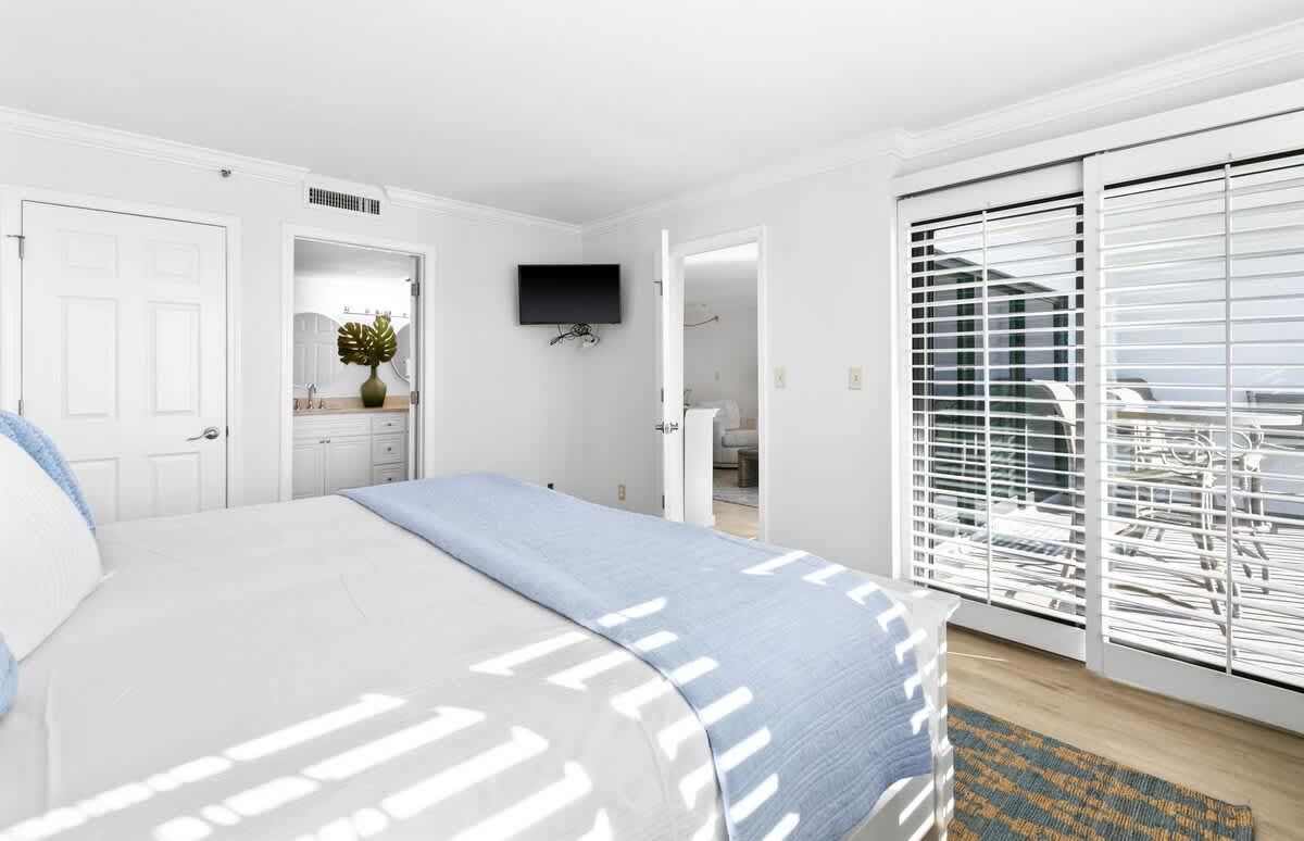 This bedroom features a white king-sized bed with blue and white bedding, a wall-mounted TV, and sliding glass doors with white plantation shutters leading to a private balcony with ocean views.