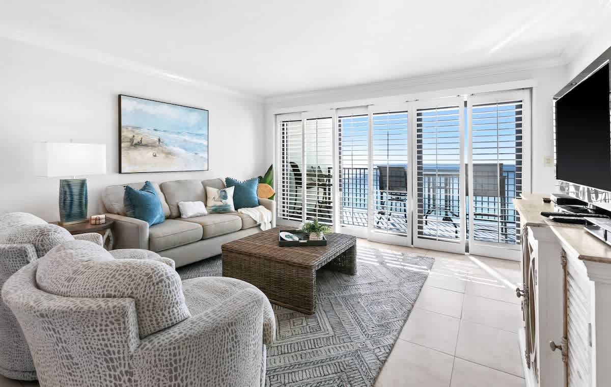 Inviting living room with a light gray sofa, two patterned armchairs, a woven coffee table, and a large area rug, offering panoramic ocean views through sliding glass doors to a balcony.