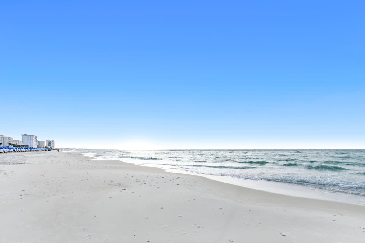 Wide, pristine white sand beach stretching along the coastline with gentle waves of the ocean. Distant buildings and blue beach umbrellas are visible under a clear blue sky.