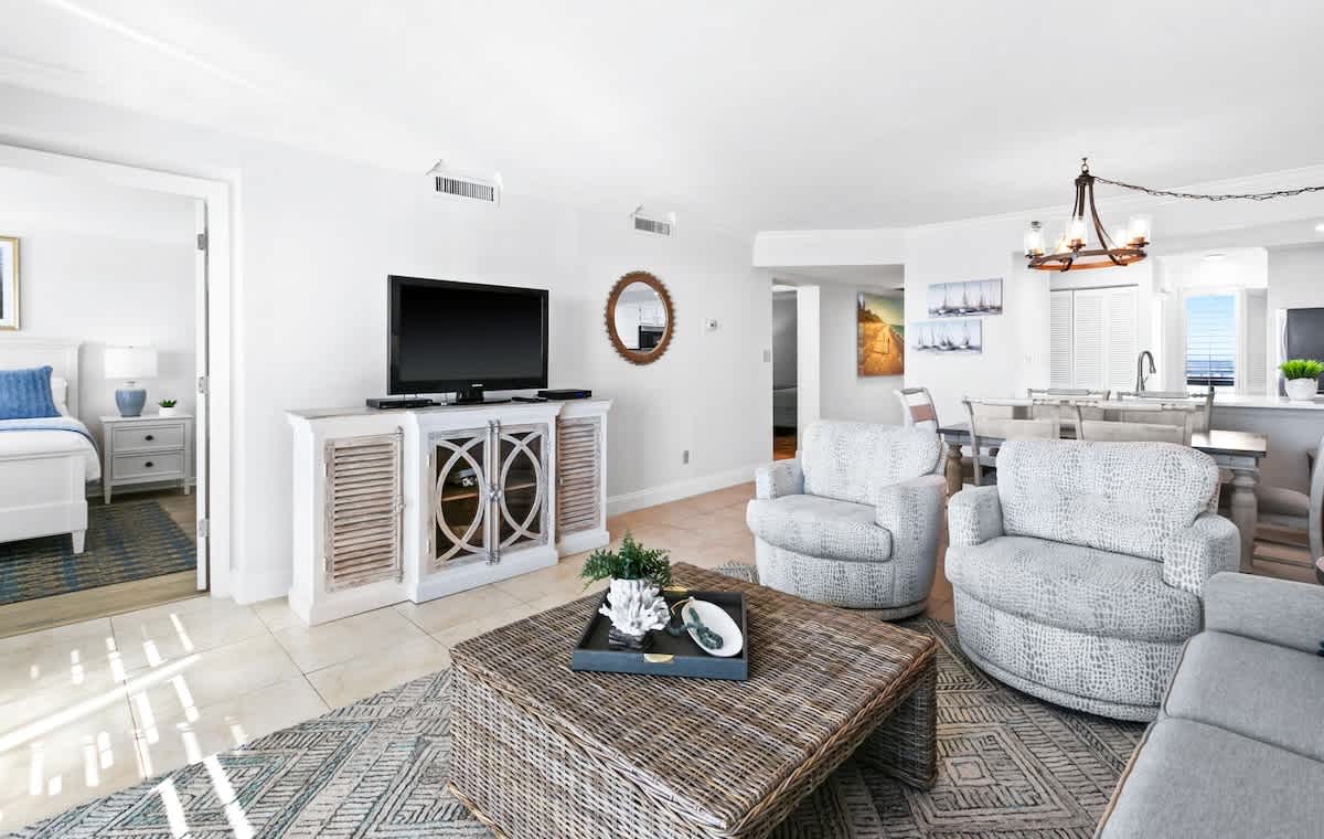 Bright living room with light tile flooring, a large flat-screen TV on a white console, two swivel armchairs, and a woven coffee table, leading to an open dining area and kitchen.