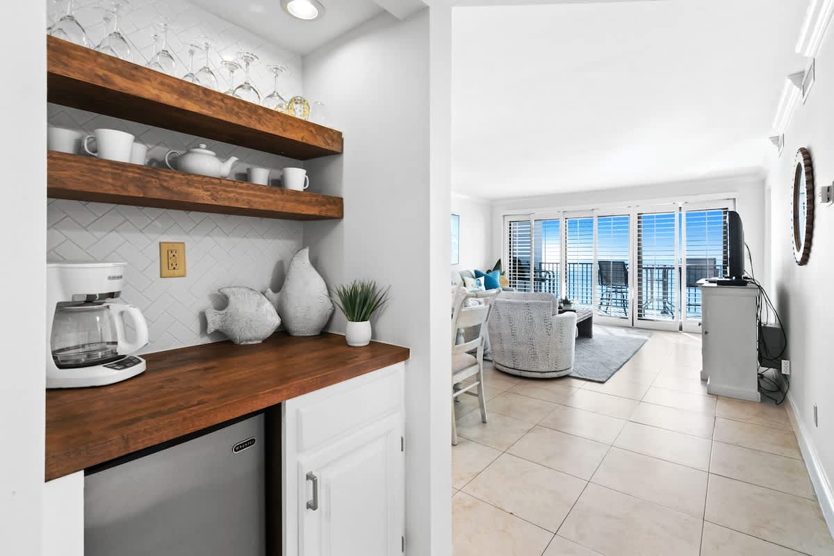 Compact wet bar or coffee station with a wooden countertop, open wooden shelving displaying glassware and dishes, a mini-fridge, and a white fish-scale tile backsplash.