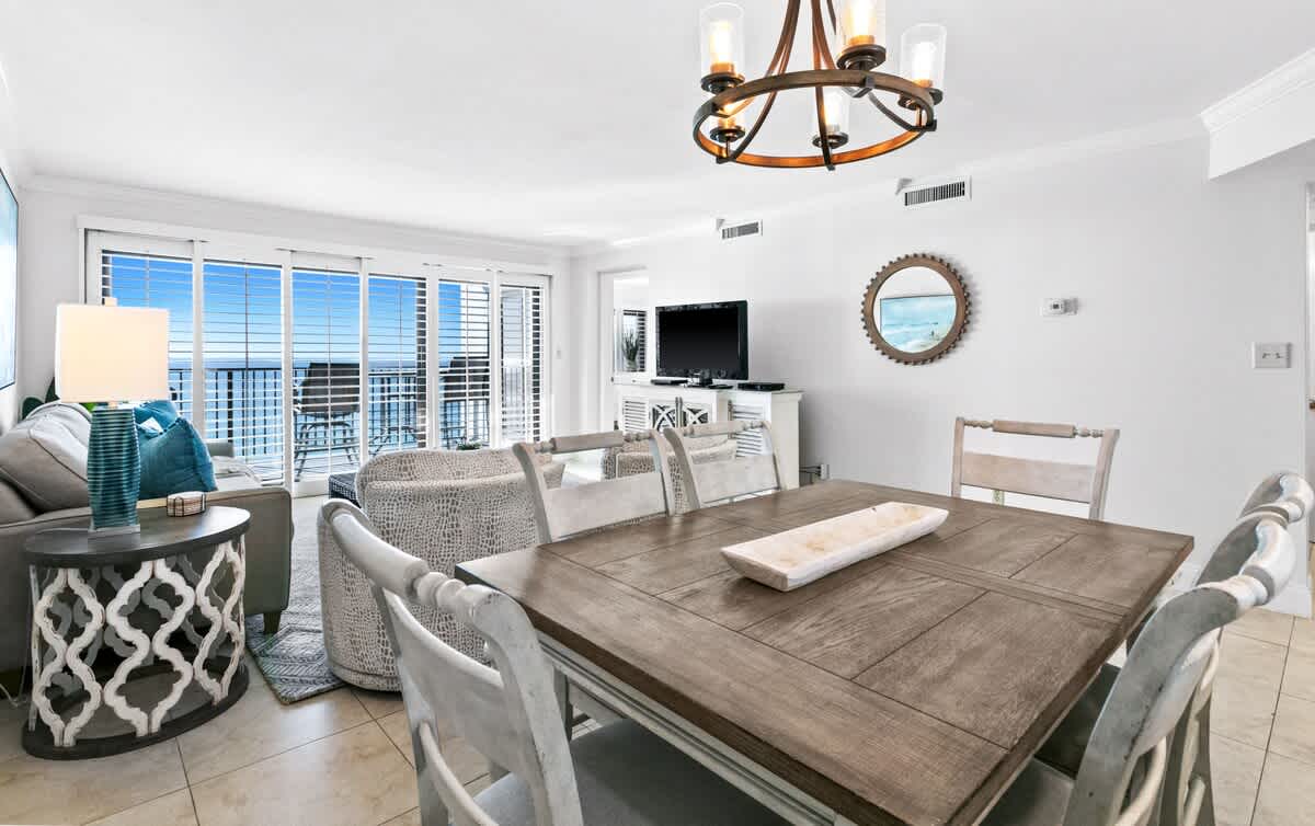 A bright, open-concept dining and living area features a rustic wooden dining table with white chairs, a gray sofa, and a blue accent lamp, all overlooking a balcony with ocean views through large sliding glass doors.