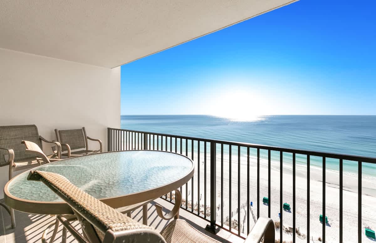 A spacious private balcony features a glass-top dining table with chairs, offering panoramic views of the bright blue ocean and white sand beach under a clear sky.