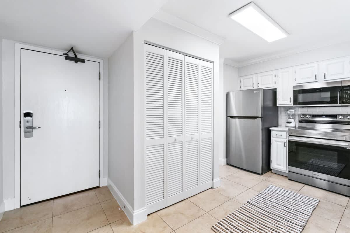 Entryway with a white front door and tiled floor, leading into a kitchen area with white cabinets and stainless steel appliances, featuring louvered bifold closet doors.