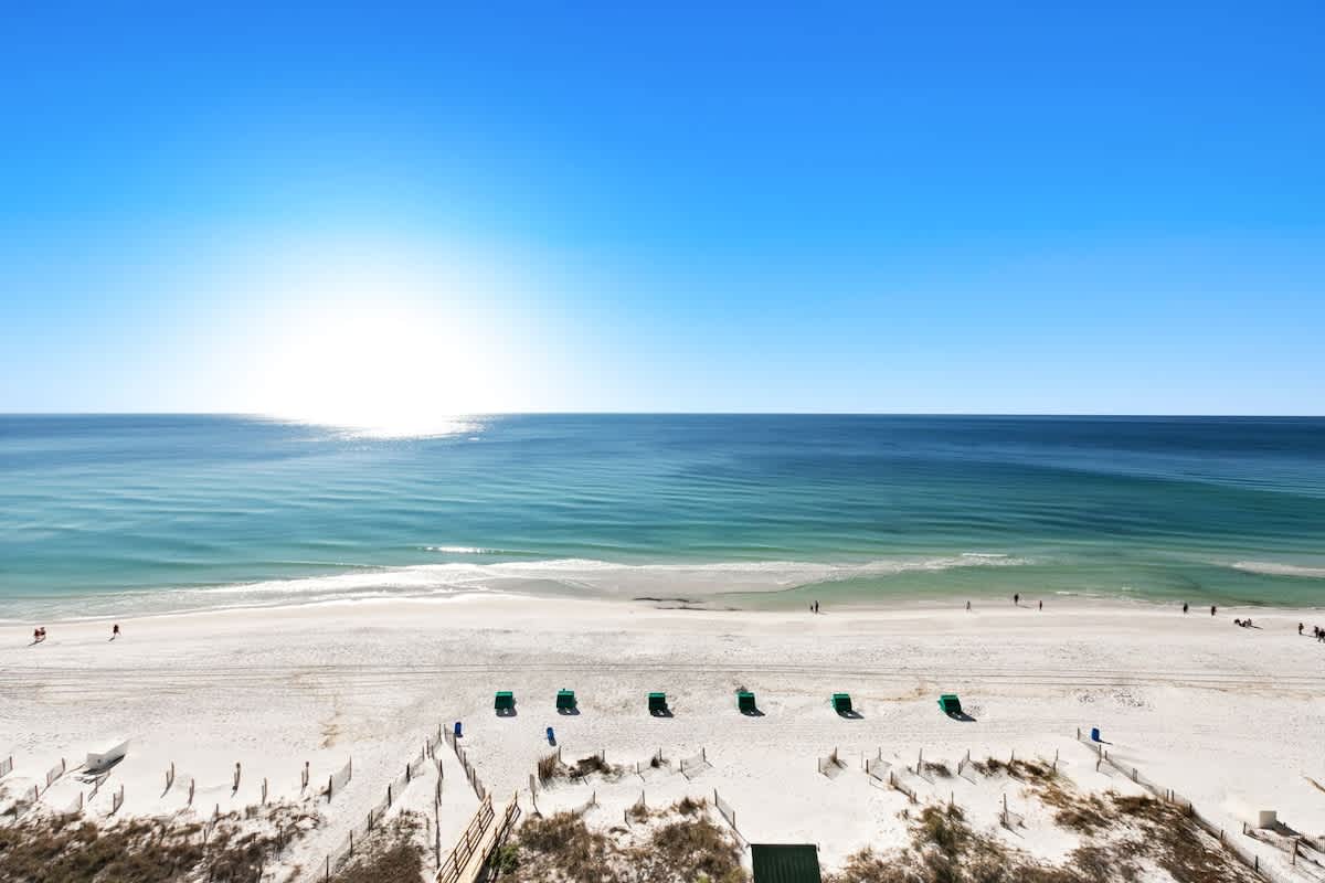 A stunning aerial view captures the expansive white sand beach and the clear turquoise ocean under a bright blue sky, with beach chairs and umbrellas dotting the shoreline.