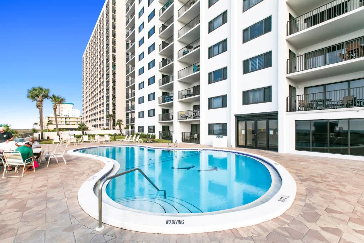 Close-up view of a freeform communal swimming pool surrounded by a paved deck. The white condominium building with multiple balconies rises behind the pool area.
