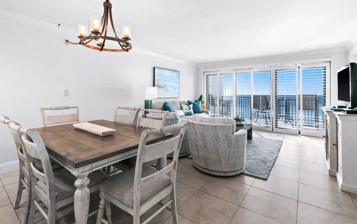 The open living and dining space features a rustic wooden dining table with white chairs, a gray sofa with blue pillows, and two patterned swivel chairs, all facing sliding glass doors that open to an ocean-view balcony.