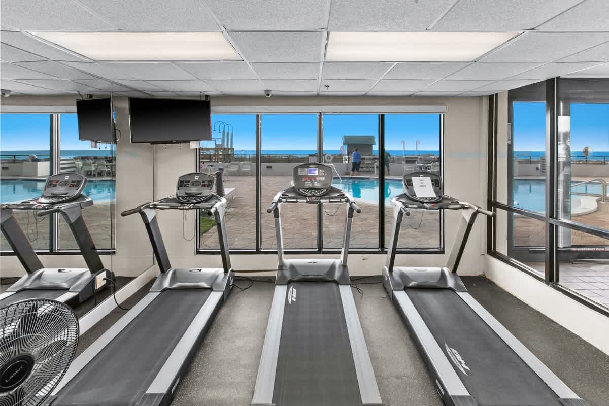 Fitness center with several treadmills facing large windows that offer views of the outdoor pool area and the ocean beyond. Flat-screen TVs are mounted above the treadmills.