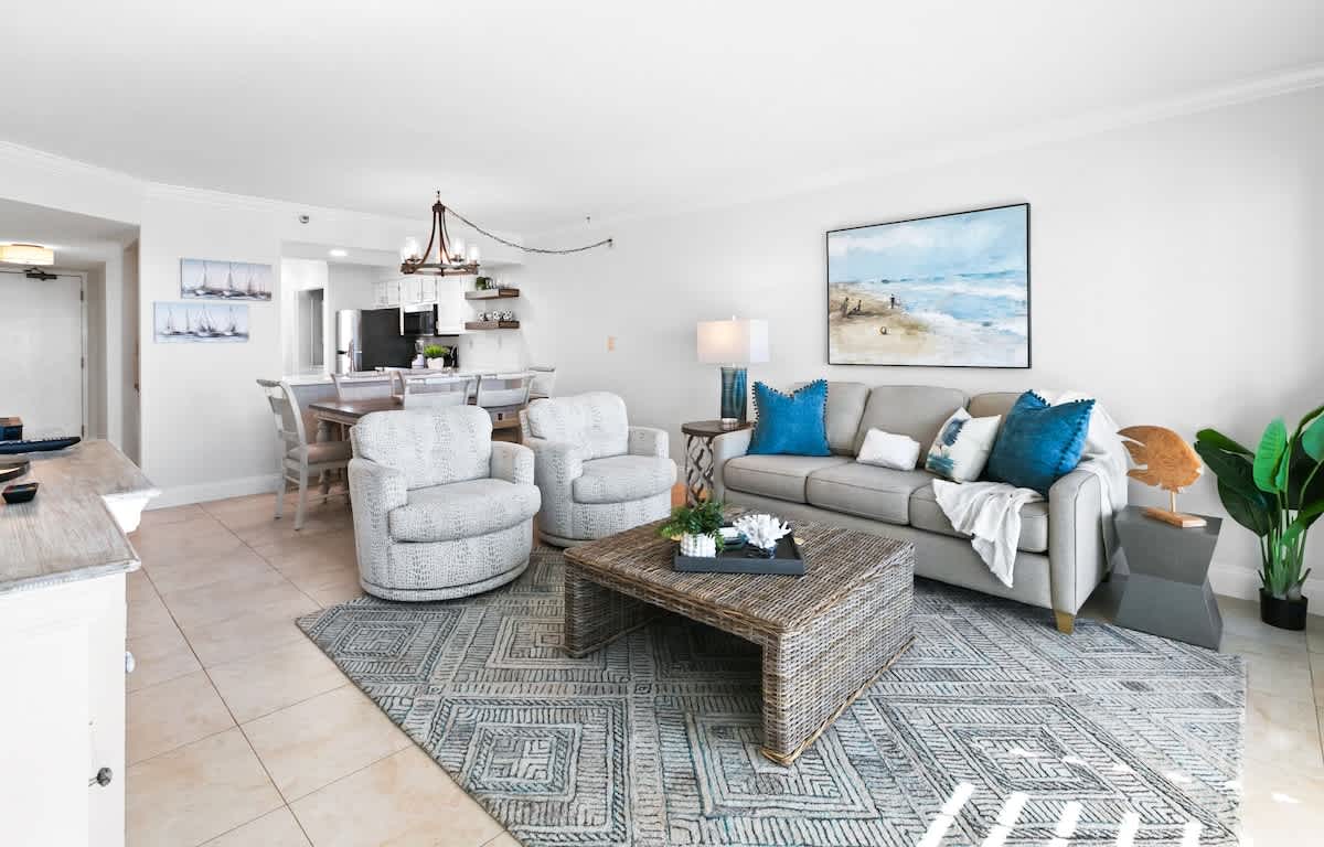 A cozy living room features a gray sofa with teal and white throw pillows, two patterned swivel chairs, and a woven coffee table on a geometric area rug, with a large ocean-themed painting on the wall.