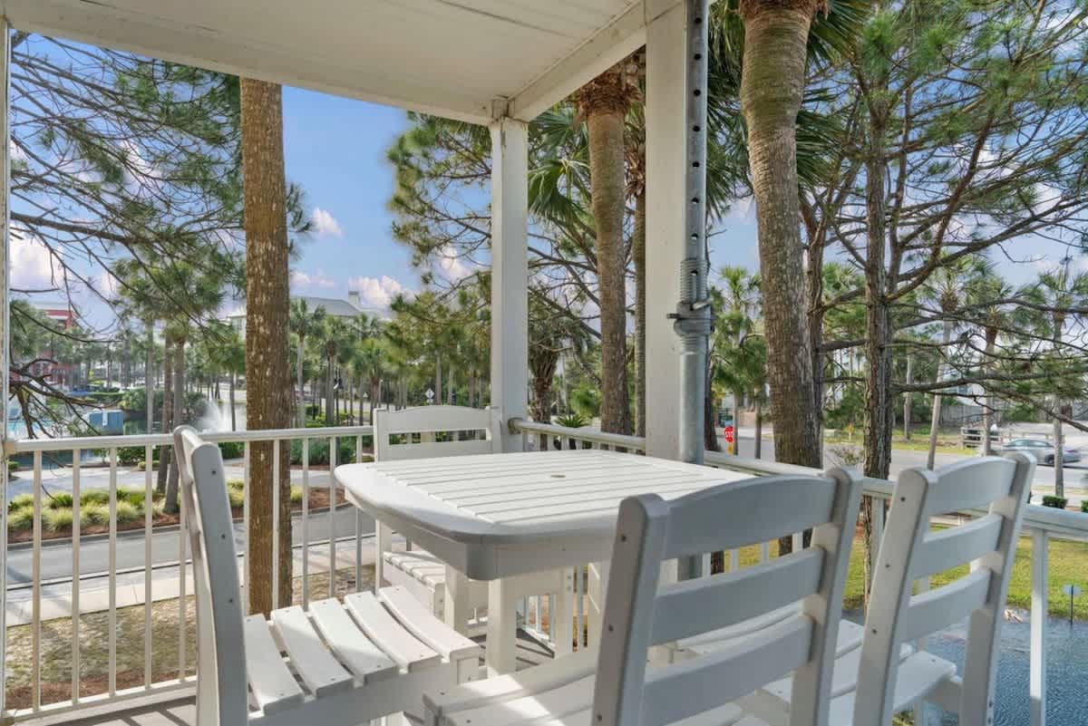 A spacious balcony is furnished with a white square dining table and four matching chairs, surrounded by dense green foliage and tall palm trees, with a road visible in the background.