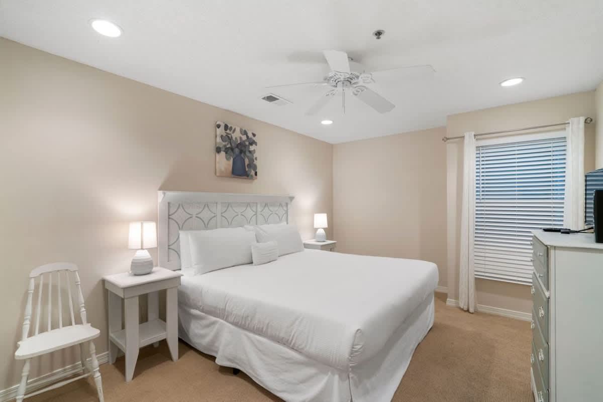 The bedroom is furnished with a white patterned headboard and a large bed dressed in white, flanked by white nightstands with lamps, and a white dresser with a TV, all on beige carpet.