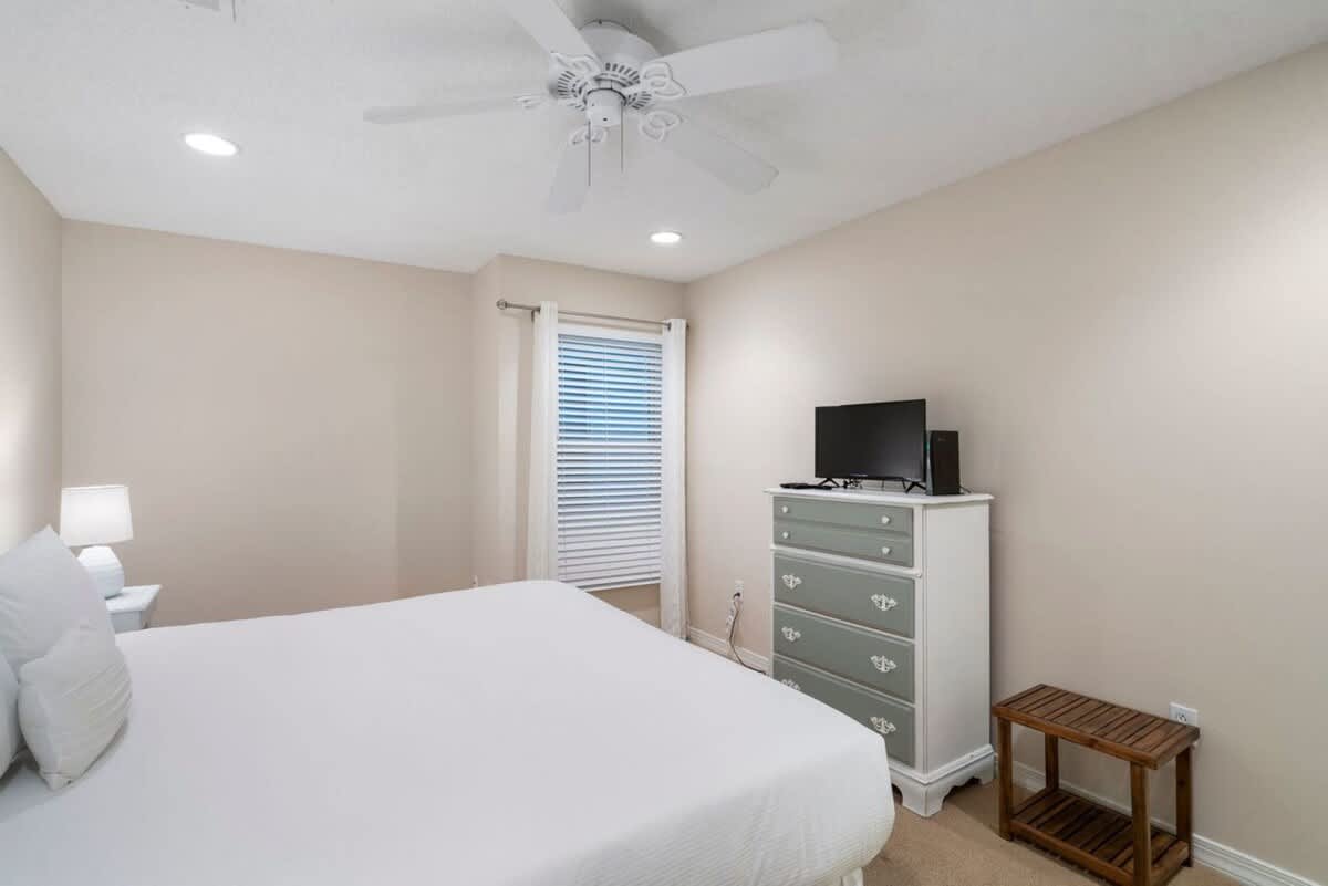A bright bedroom showcases a large bed with white bedding, a white dresser with light green drawers and a TV, and a window with white blinds, complemented by light beige walls and carpet.