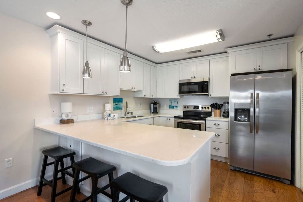 Bright kitchen featuring white shaker cabinets, light quartz countertops, and stainless steel appliances including a refrigerator, microwave, and range. A breakfast bar with three black stools provides casual seating.