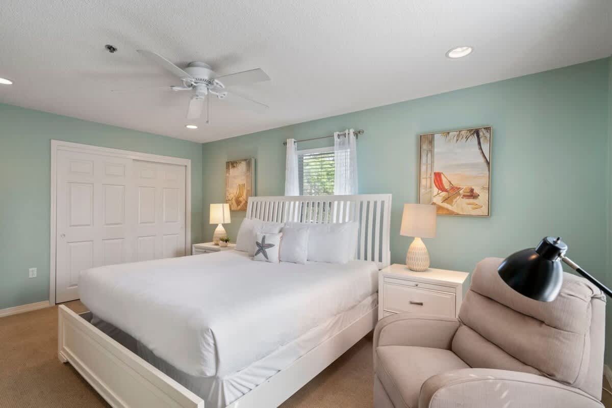 A spacious bedroom with light teal walls is furnished with a white slatted headboard on a large bed with white linens, a comfortable beige recliner, and a large closet with white bi-fold doors.
