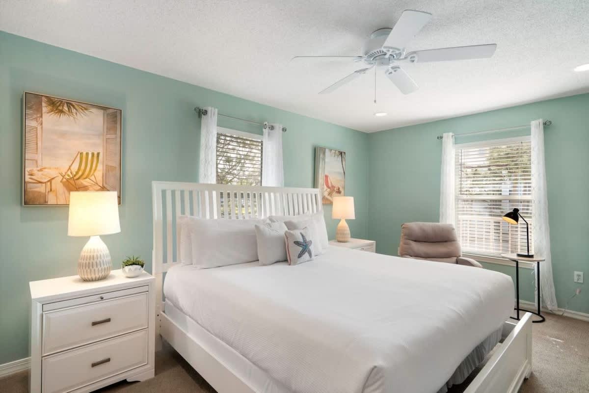 This bright bedroom features light teal walls, a white slatted headboard on a large bed with white linens, two windows with white curtains, and white nightstands with lamps, creating a serene atmosphere.