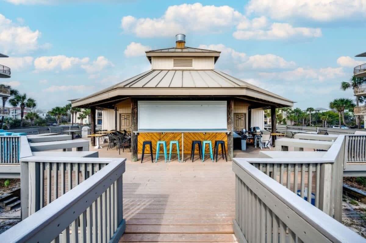 Elevated wooden pavilion featuring an outdoor bar with stools, surrounded by railings and offering views towards the beach, under a bright blue sky with scattered clouds.